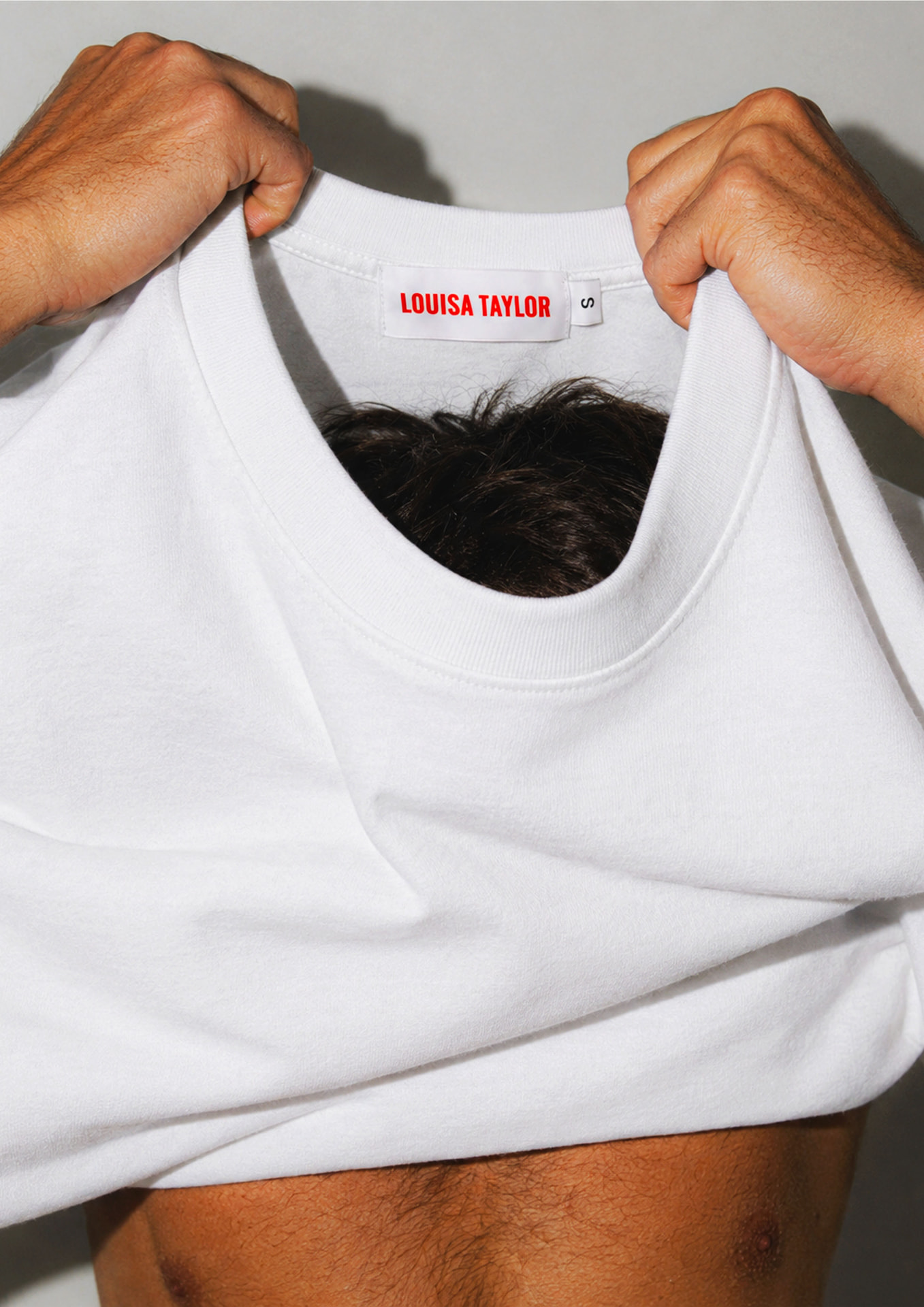 Hands pulling a white tee over the head, revealing the red Louisa Taylor neck label